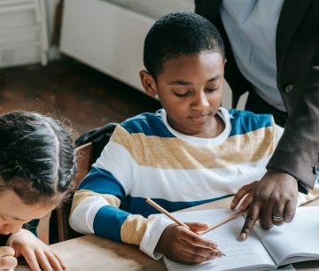 High angle of crop unrecognizable black female teacher explaining task to focused little schoolboy sitting at desk near attentive Asian classmate writing in notebook